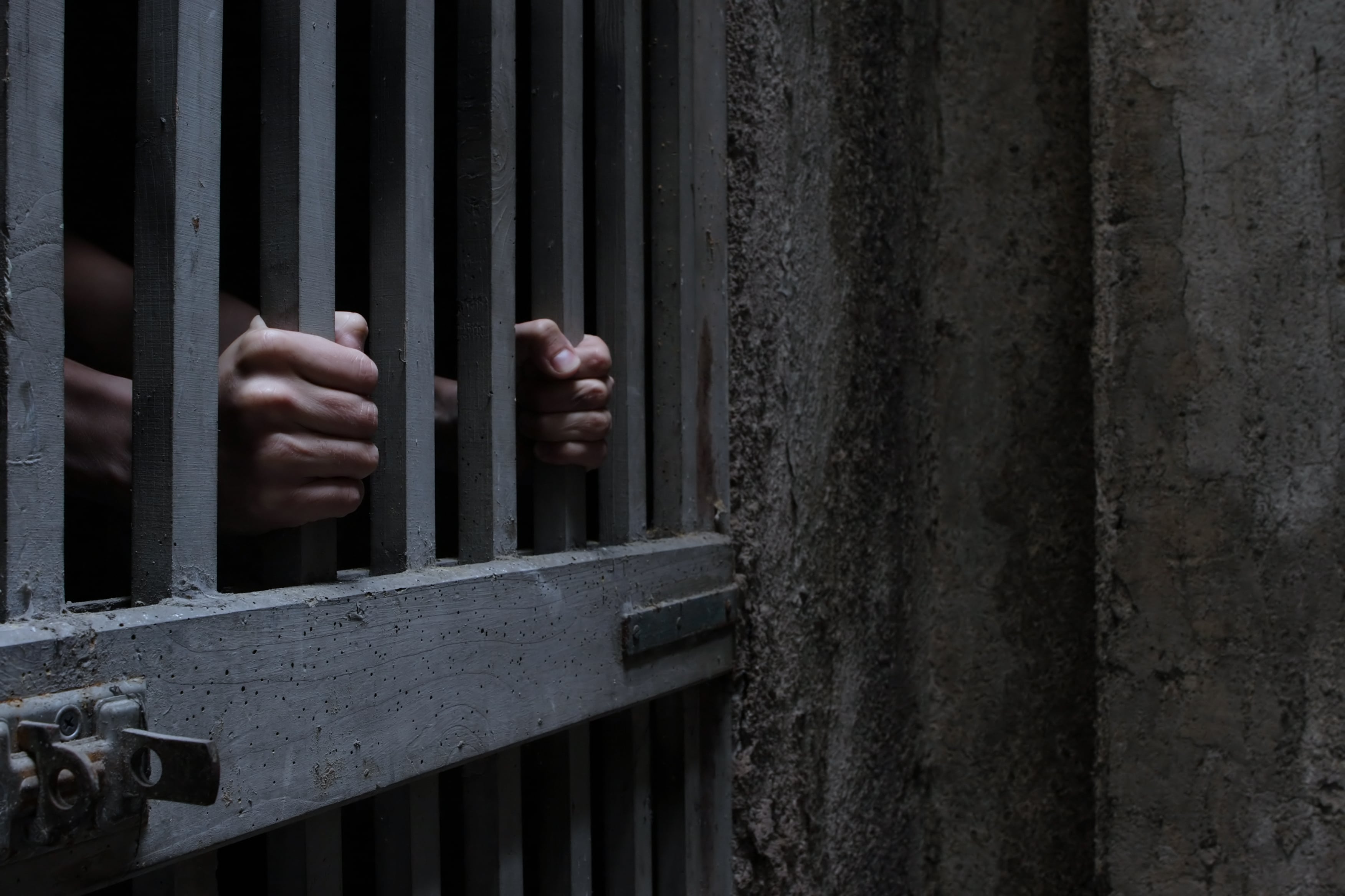Hand making a peace sign through bars of a white, weathered prison cell, with another hand visible, suggesting confinement and hope.