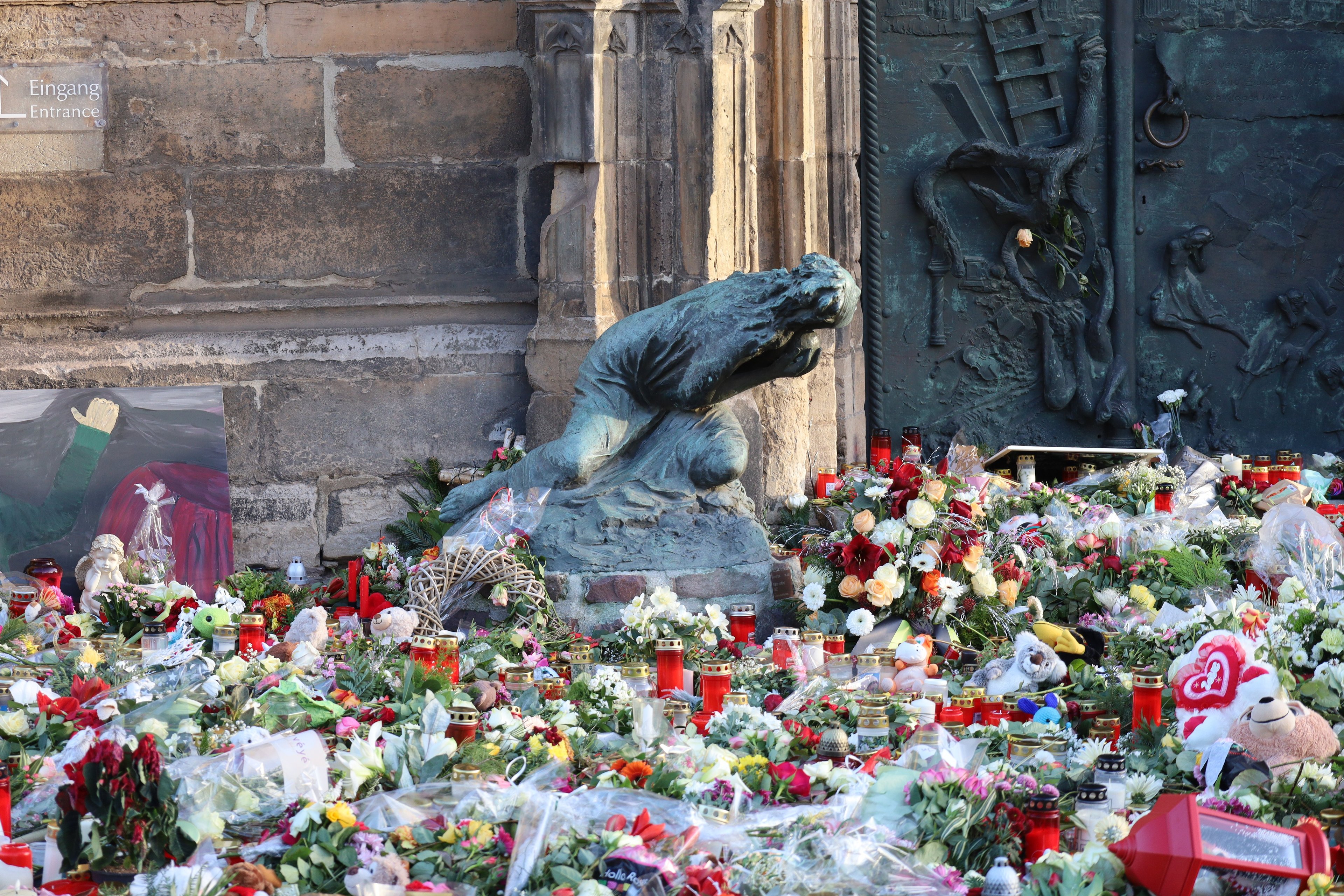 A memorial site with numerous flower bouquets and candles on the ground, surrounded by a crowd of people paying their respects.