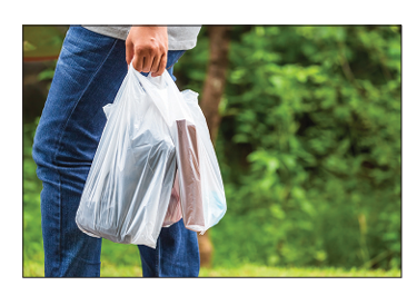 Photograph of a person's hand carrying two white plastic carrier bags outdoors, with green foliage in the background