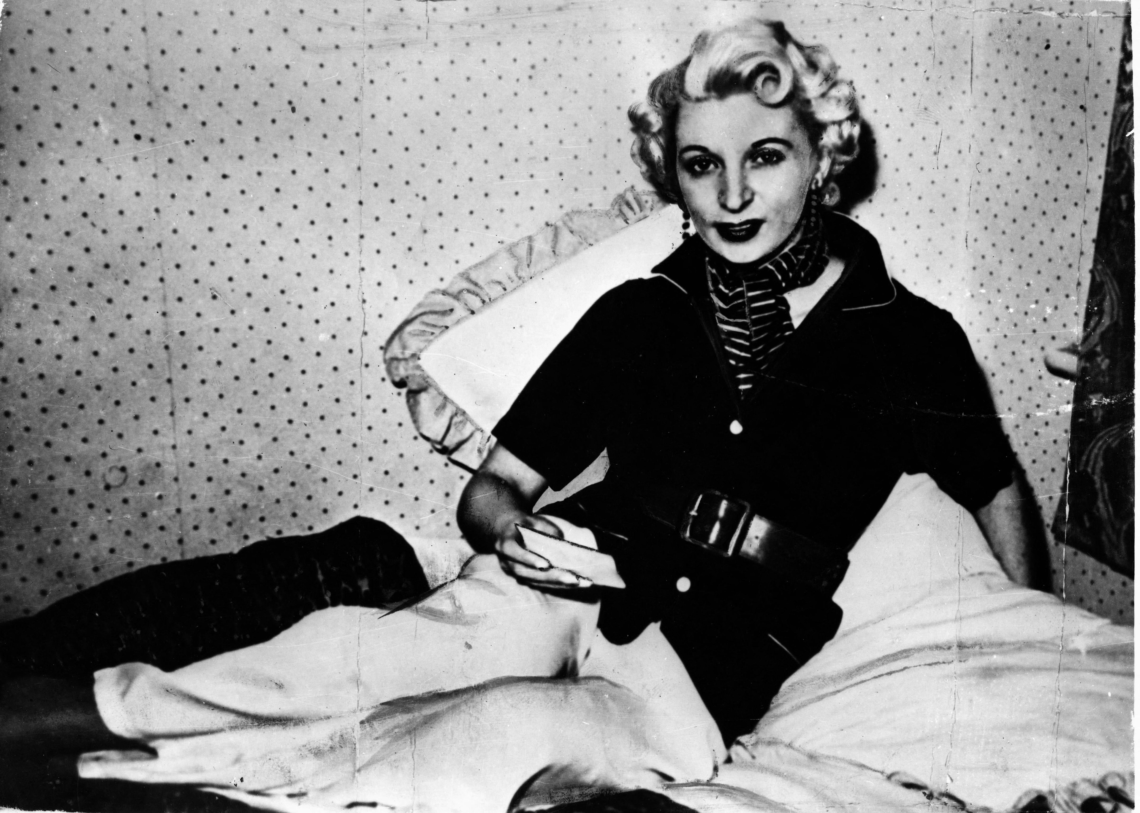 Black-and-white portrait of a stylish woman with short curled blonde hair, dark lipstick, striped scarf and earrings, posing before a patterned backdrop