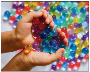 Photograph of two hands cupping a handful of small, translucent hydrogel beads in red, blue, green, yellow and purple, with many more colourful beads scattered across the background.