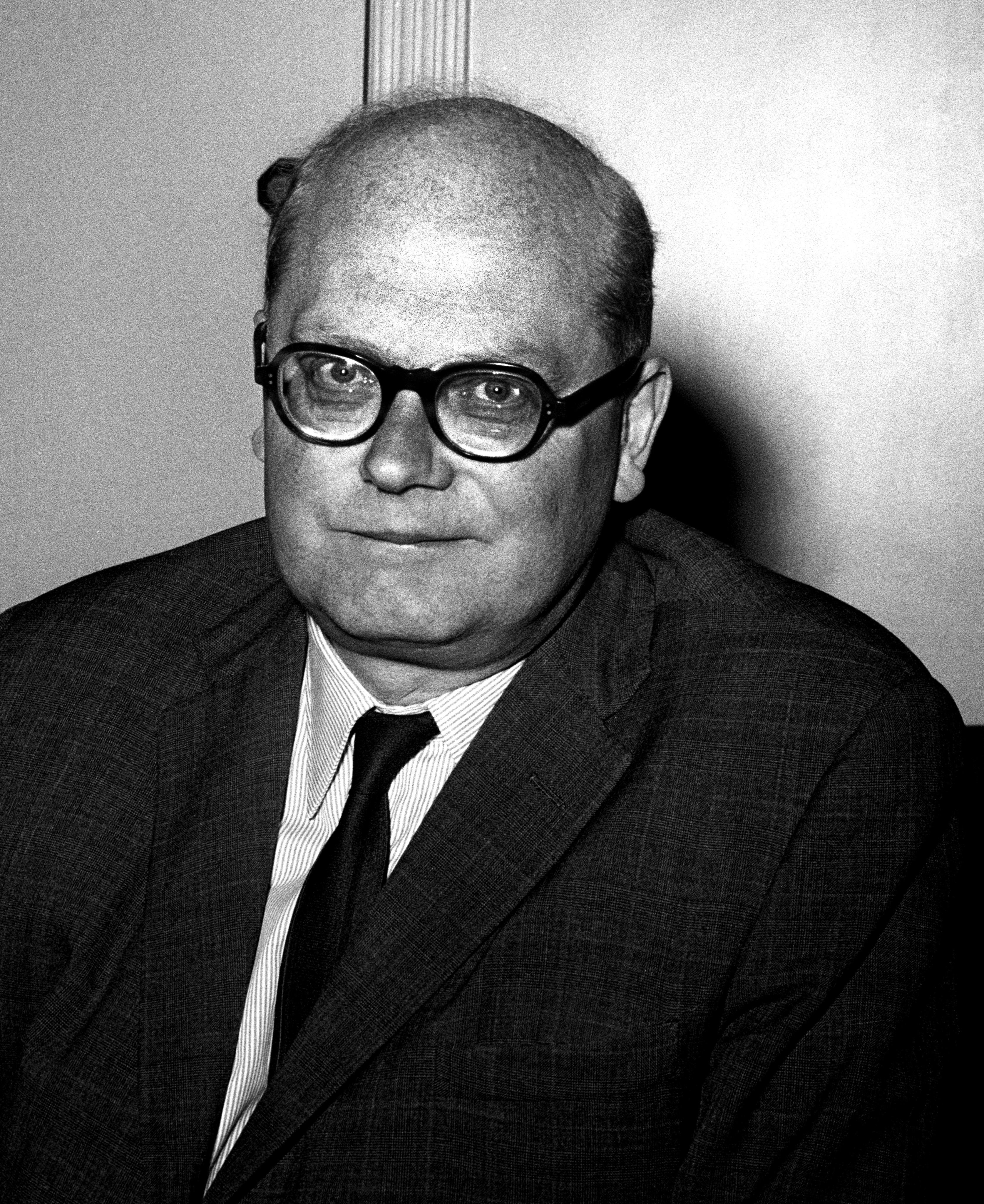Black-and-white portrait of an older man in glasses and suit, smiling slightly while looking past the camera, against a softly blurred dark background.