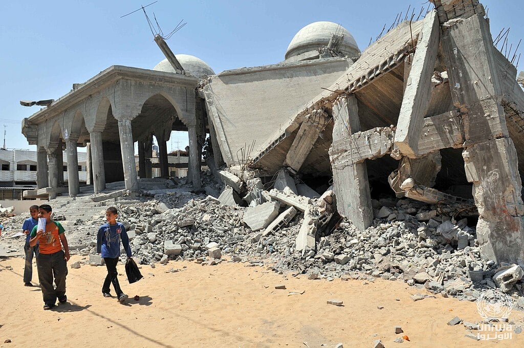 A woman carrying a child and holding a girl's hand walks through rubble and debris, with more children in the background, in an area affected by conflict.