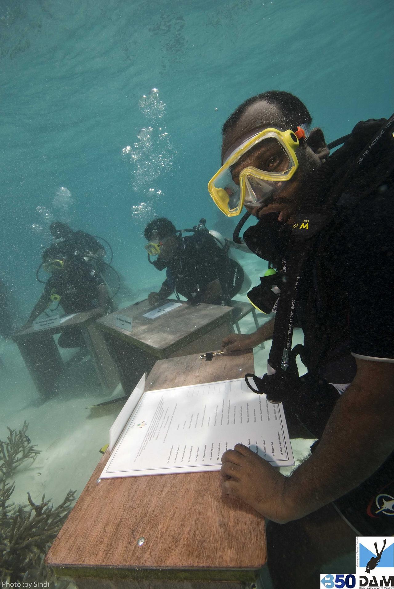 Scuba divers in wetsuits and masks sign documents at an underwater table, with name cards visible, in a unique aquatic setting.