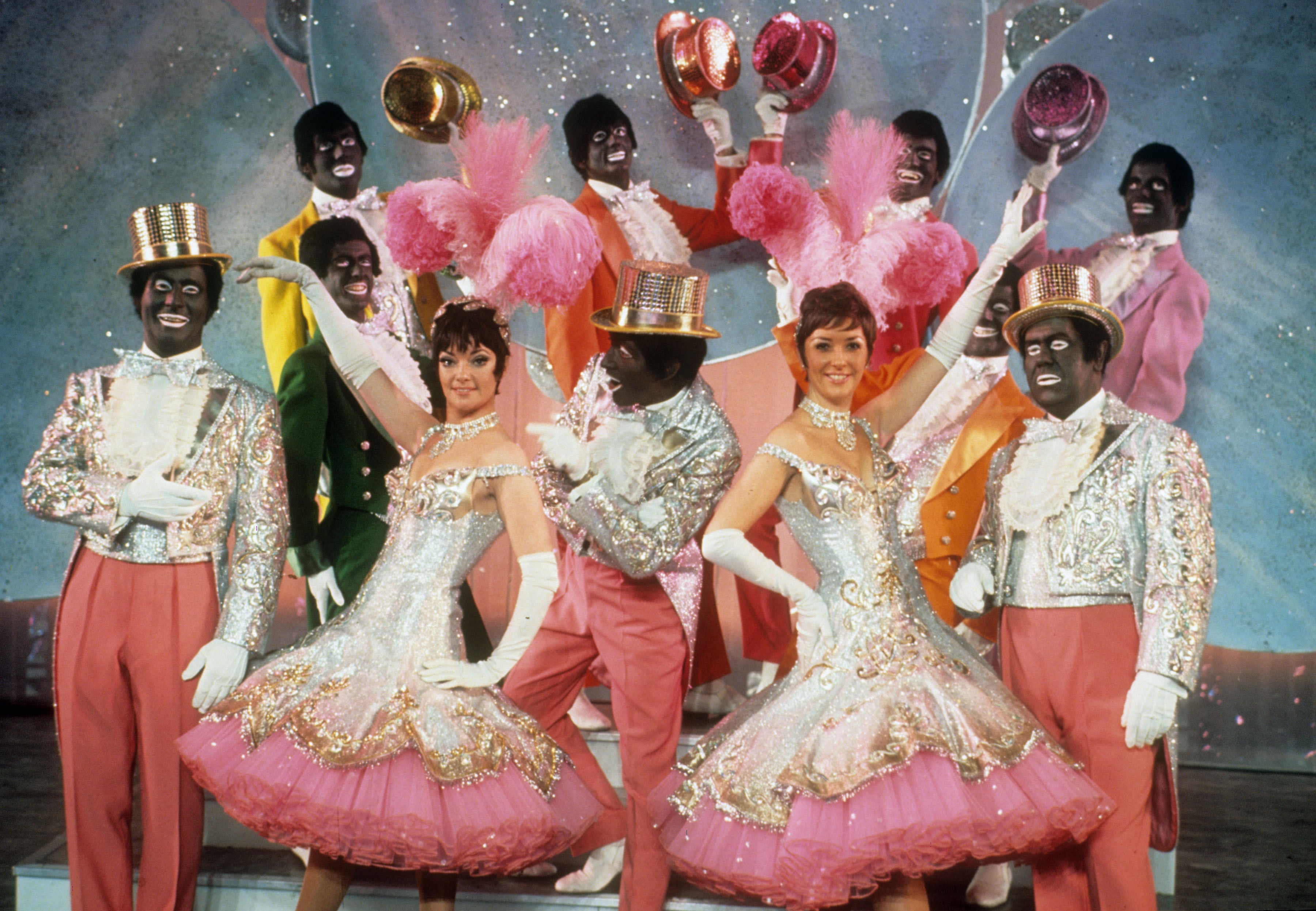 Stage performers in glittering white tailcoats and top hats wearing blackface makeup, singing and dancing in a choreographed musical routine
