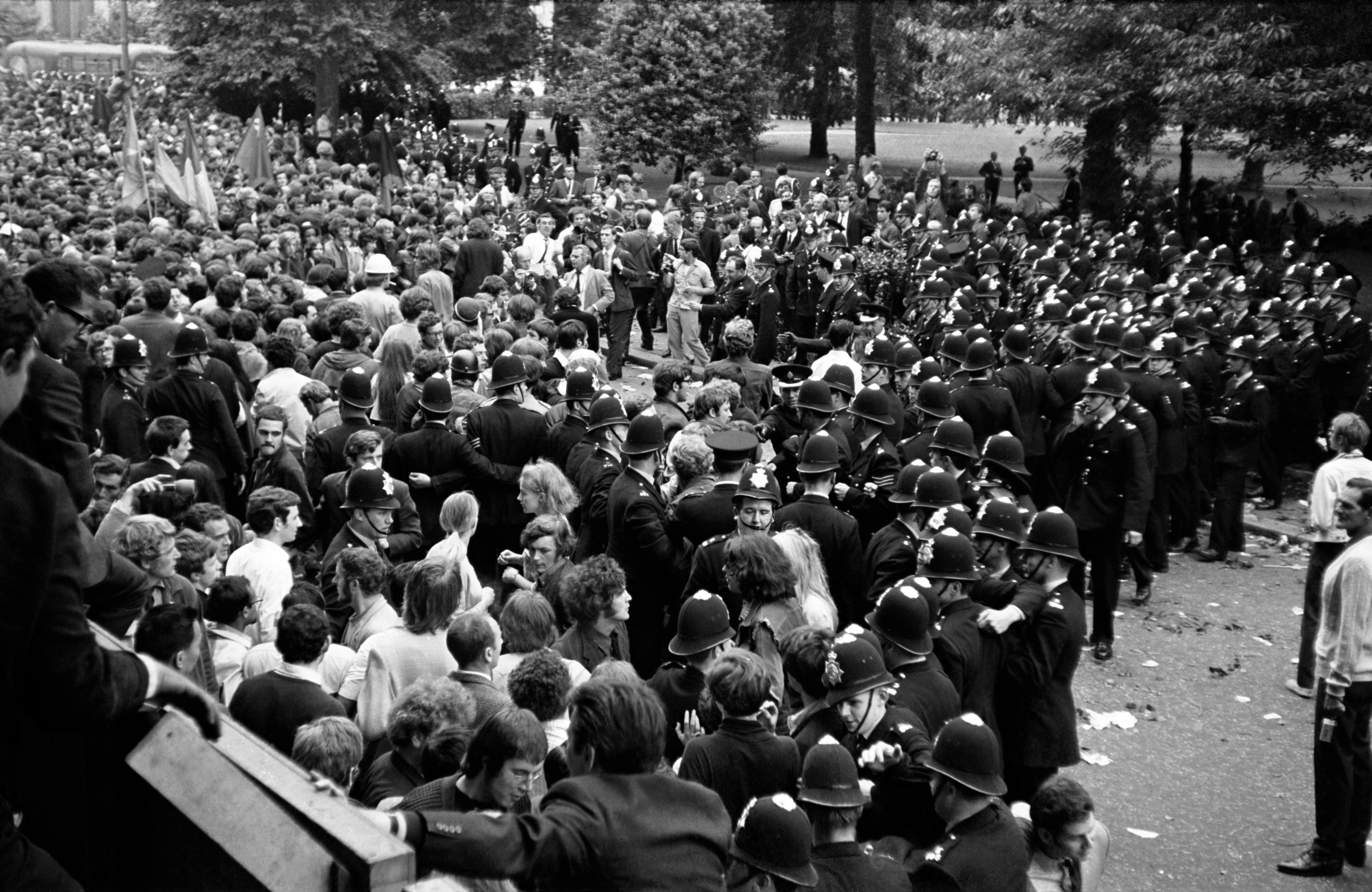 Large black-and-white crowd scene with mounted police containing protesters in a park, officers lining the road and scattered debris on the ground