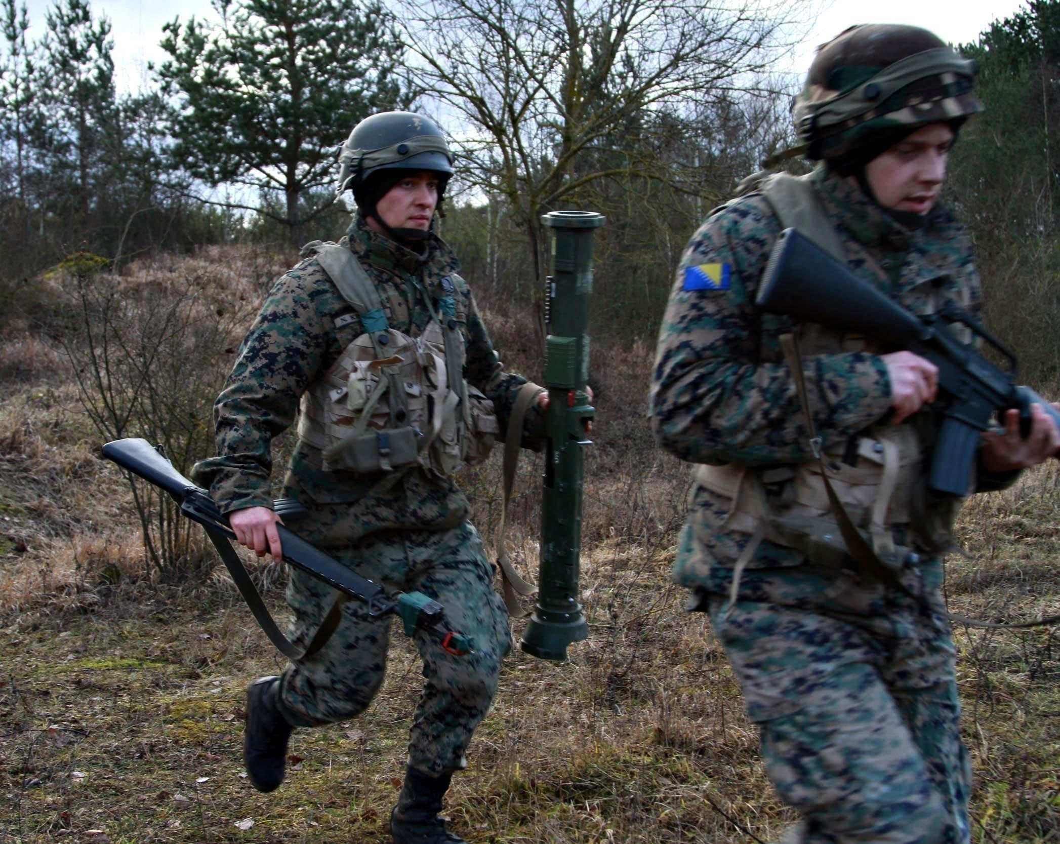 Two soldiers in camouflage uniforms, helmets and tactical vests move through a wooded area carrying rifles; the nearer soldier is blurred and the other carries a large green cylindrical weapon or launcher.