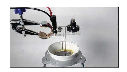 Photograph of a small electrolysis set-up — two carbon electrodes clamped and dipped into a white dish containing zinc chloride, connected by wires to a power supply with a small bulb in the circuit to show current is flowing
