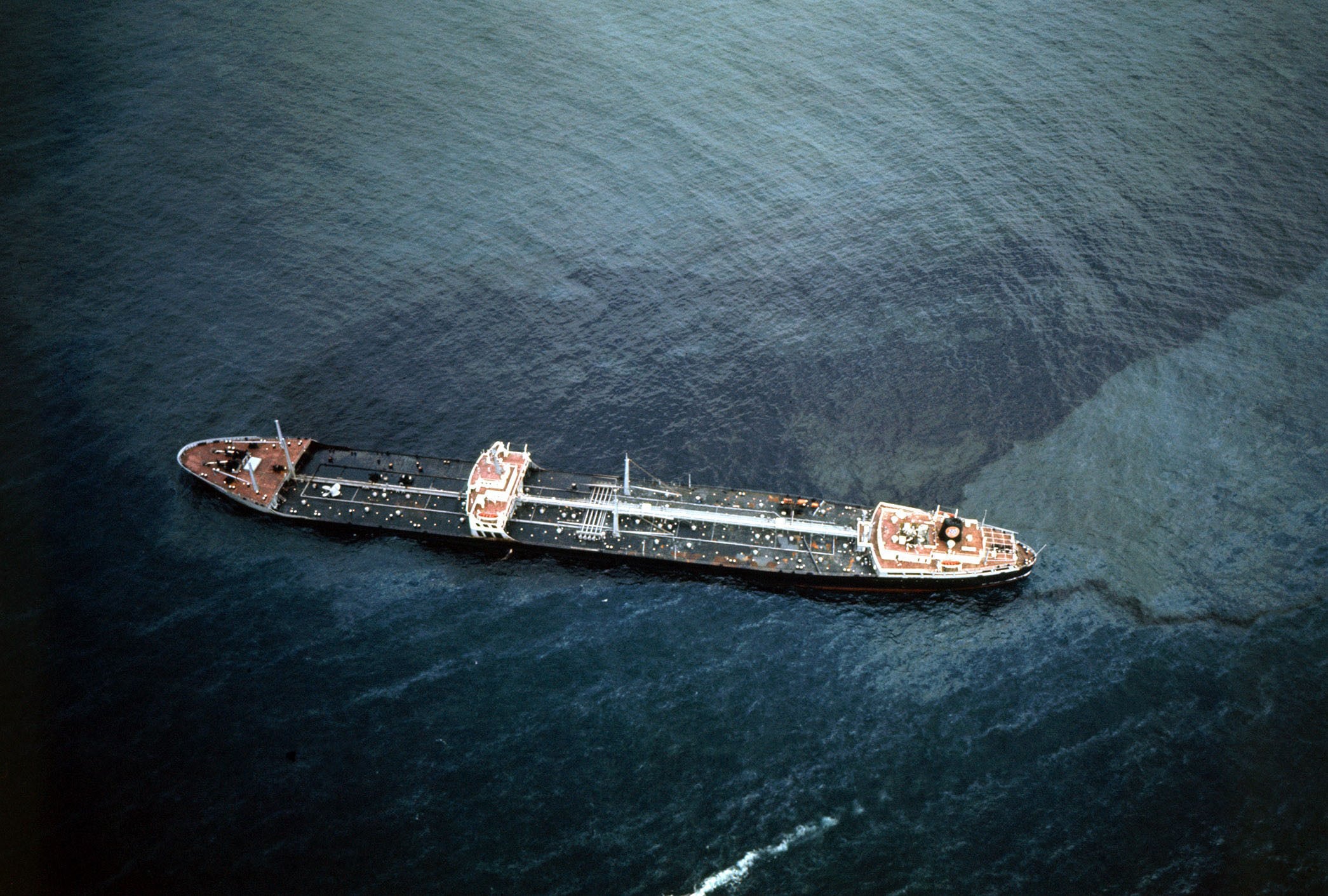 A damaged oil tanker listing in turquoise sea, surrounded by a large dark oil slick spreading across the water’s surface.