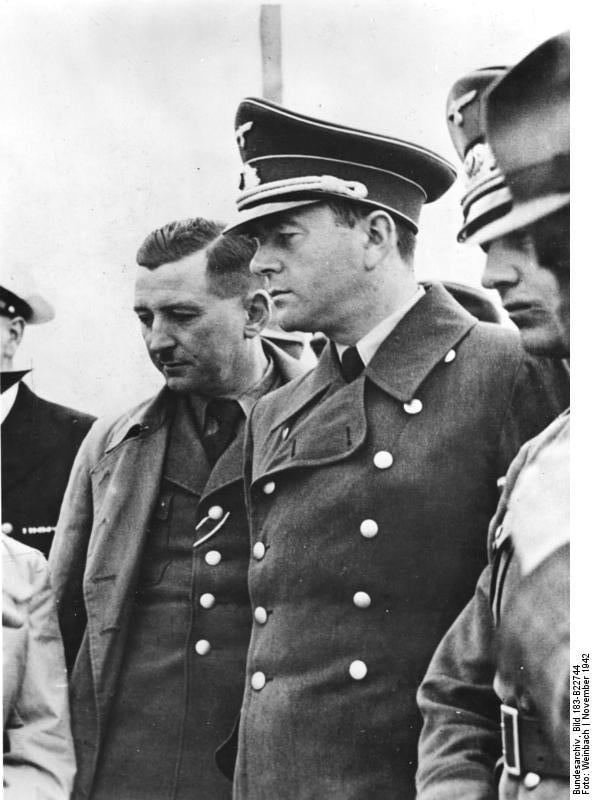 Black-and-white photograph of Albert Speer in uniform and peaked cap at the centre, standing among German officials, with Adolf Hitler partly visible behind him on the left.