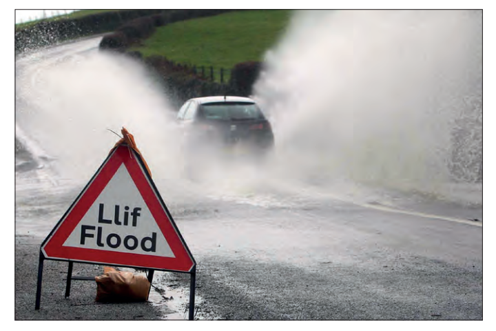Photograph of a flooded road with a car driving through deep spray; a triangular warning sign in the foreground reads “Llif Flood”.
