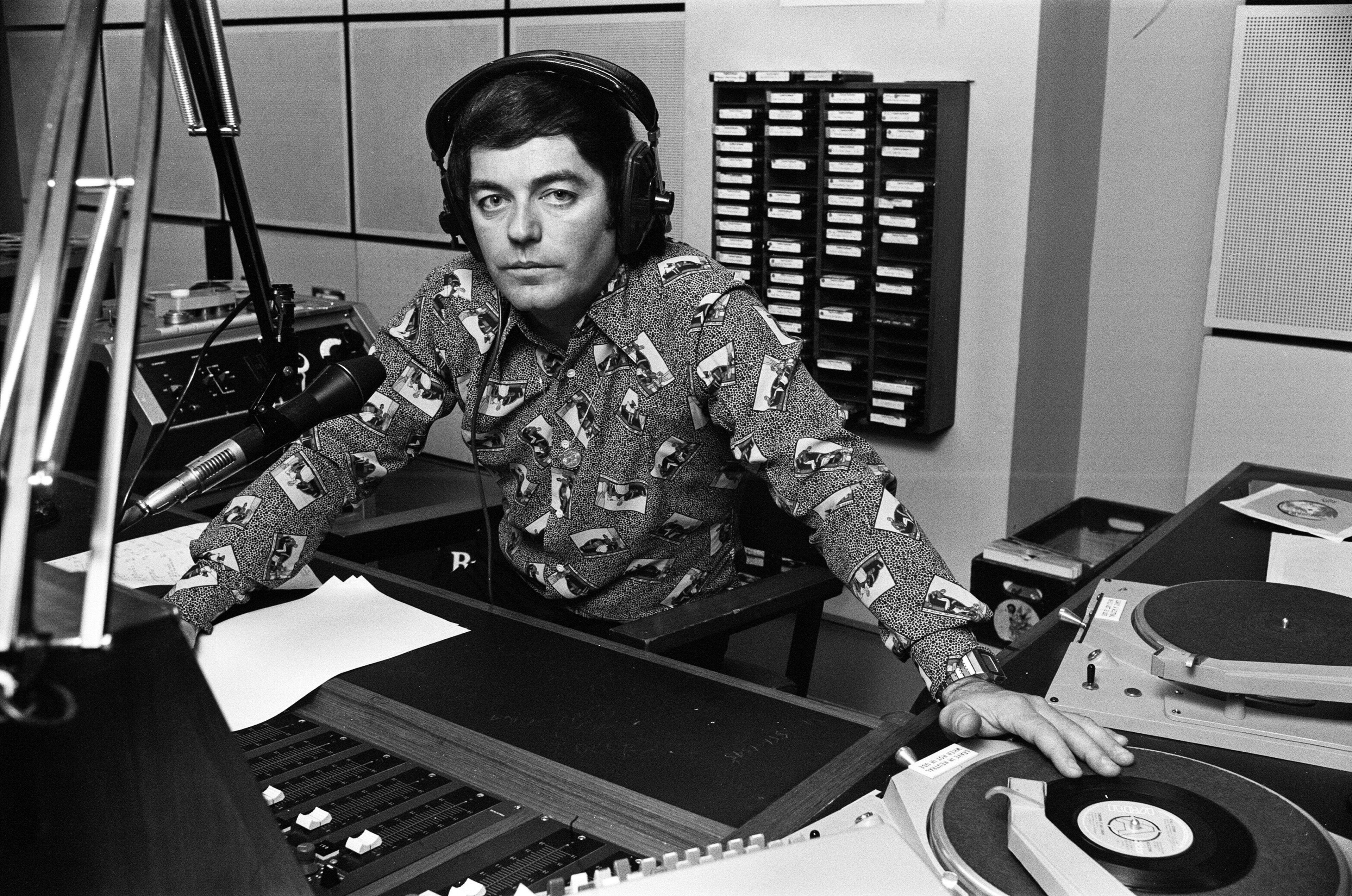 Young radio DJ in a vintage studio, smiling with headphones round his neck and a vinyl record balanced playfully on his head near mixing equipment
