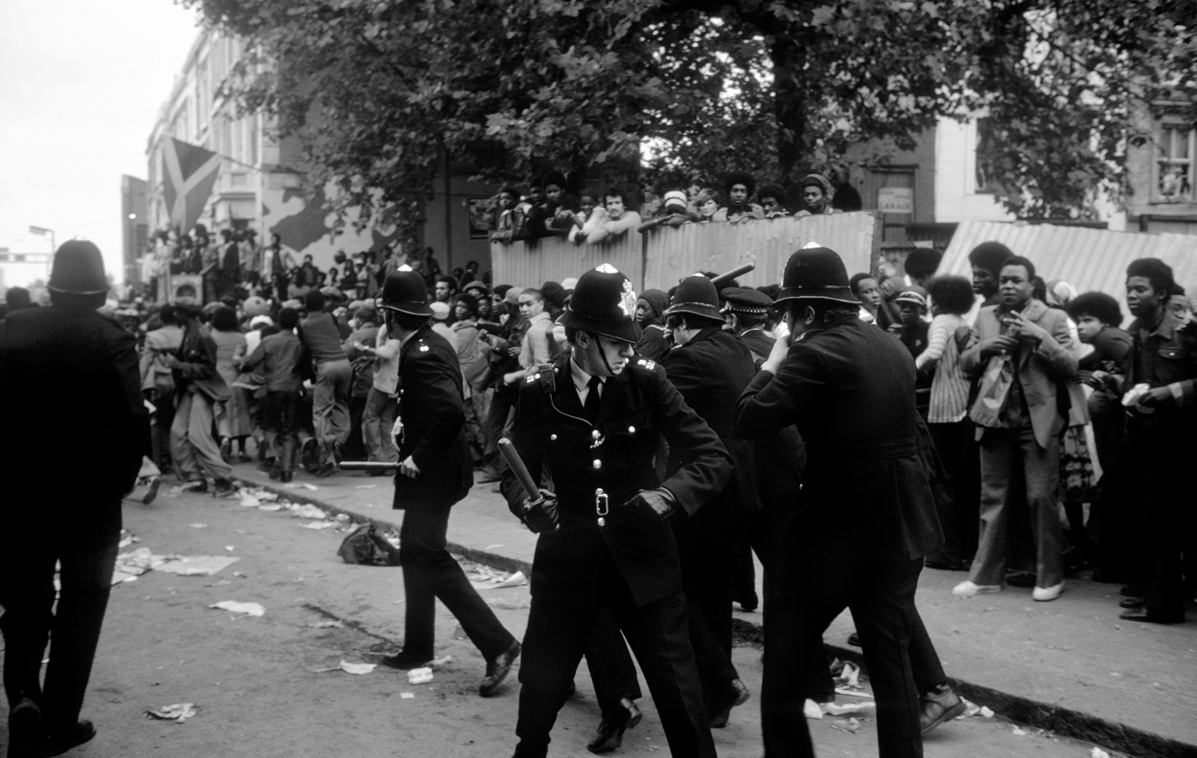 Black‑and‑white street scene of a tense clash, with a policeman grabbing a Black man amid a crowded protest or demonstration in disarray