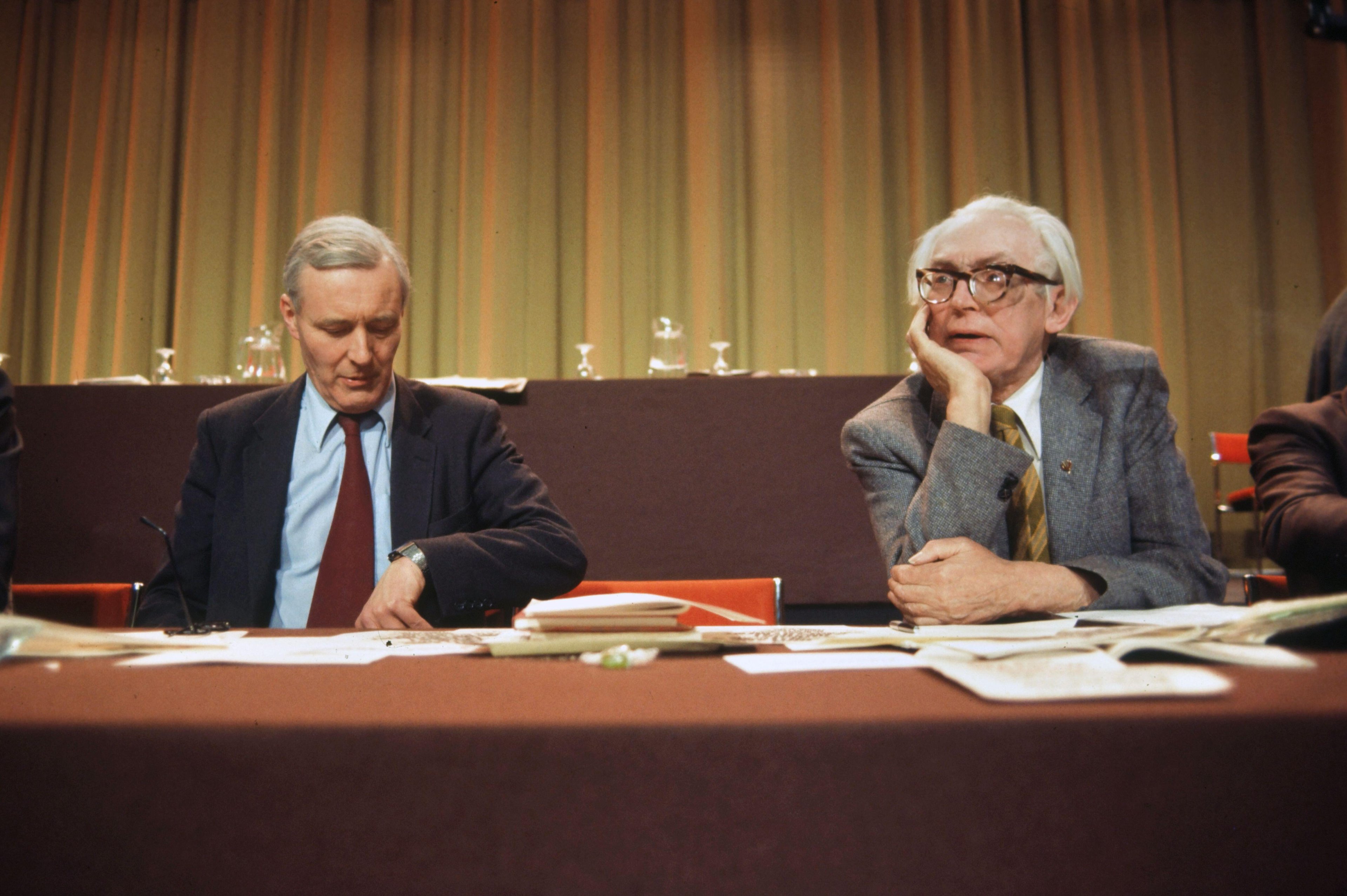 Black-and-white photo of two older men in suits seated side by side, resting chins on their hands, watching something intently in a formal setting
