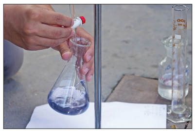 A dropper adds a few drops of indicator to the solution in a conical flask during a titration. A graduated burette containing liquid is visible beside the flask.