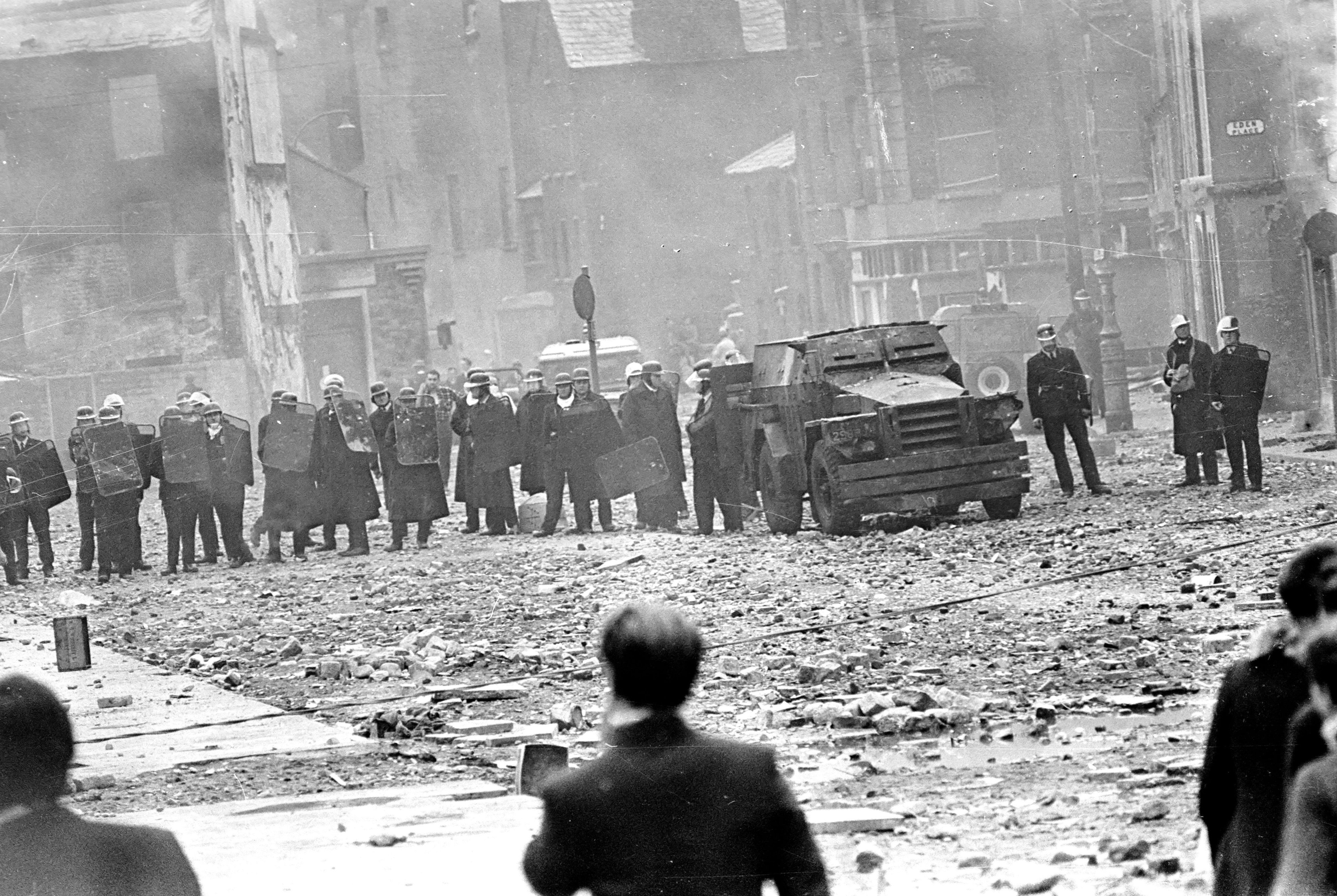 Black-and-white photograph of a line of RUC officers with riot shields and an armoured car in a rubble-strewn street, watched by onlookers