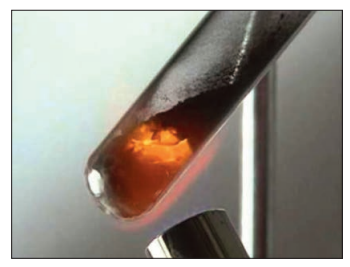 Photograph of a boiling tube containing a solid mixture glowing orange at its closed end while being heated by a Bunsen burner, illustrating the copper extraction experiment.