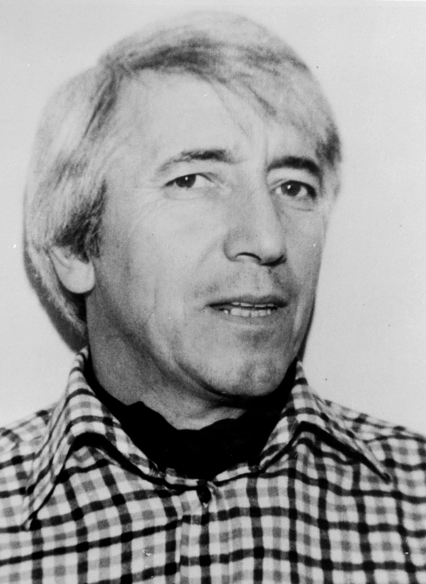 Black-and-white portrait of a middle-aged man with light hair smiling warmly, leaning slightly forward in a casual shirt against a dark background