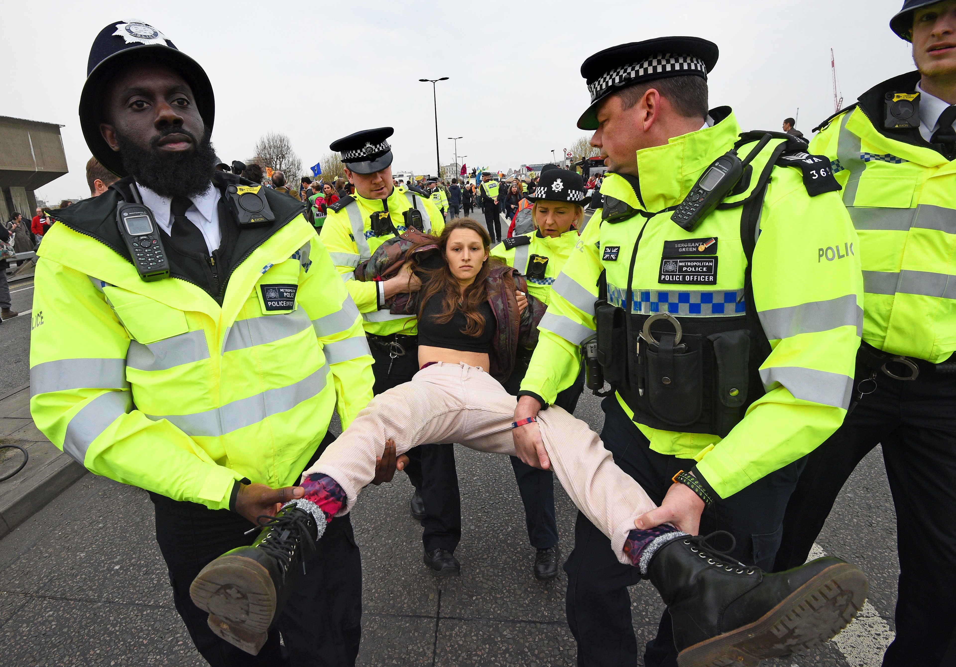 Police officers carry a woman away from a protest scene, with other officers and protesters visible in the background.