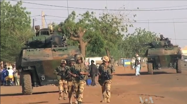 Military vehicles move through a dusty street lined with trees, accompanied by a large crowd of people, including children in colourful clothing.