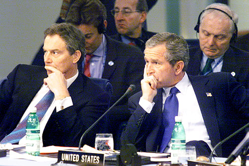 Tony Blair and George W. Bush sit side by side at an international meeting, both in suits with hands to their faces. A “UNITED STATES” nameplate and bottled water are visible on the table.