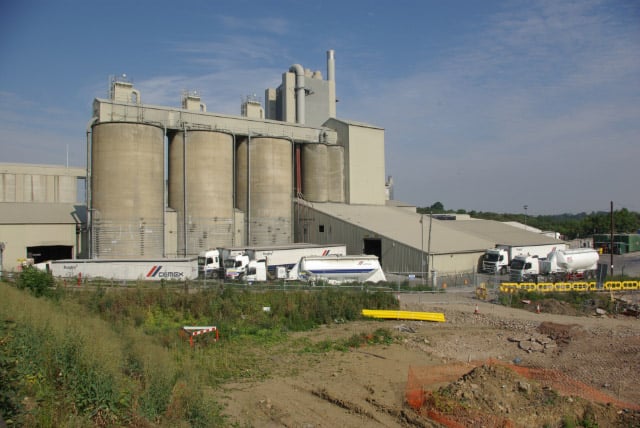 Photograph of a large cement works, showing storage silos, processing buildings and tall chimneys, with several cement tanker lorries in front and vegetation-cleared, disturbed ground around the site.