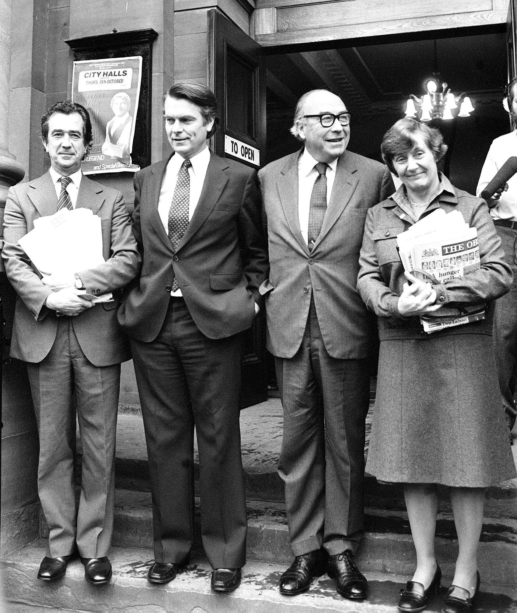 Black-and-white photograph of the Gang of Four—Bill Rodgers, David Owen, Roy Jenkins and Shirley Williams—standing outside a building in 1981, holding papers marked “SDP” and newspapers.