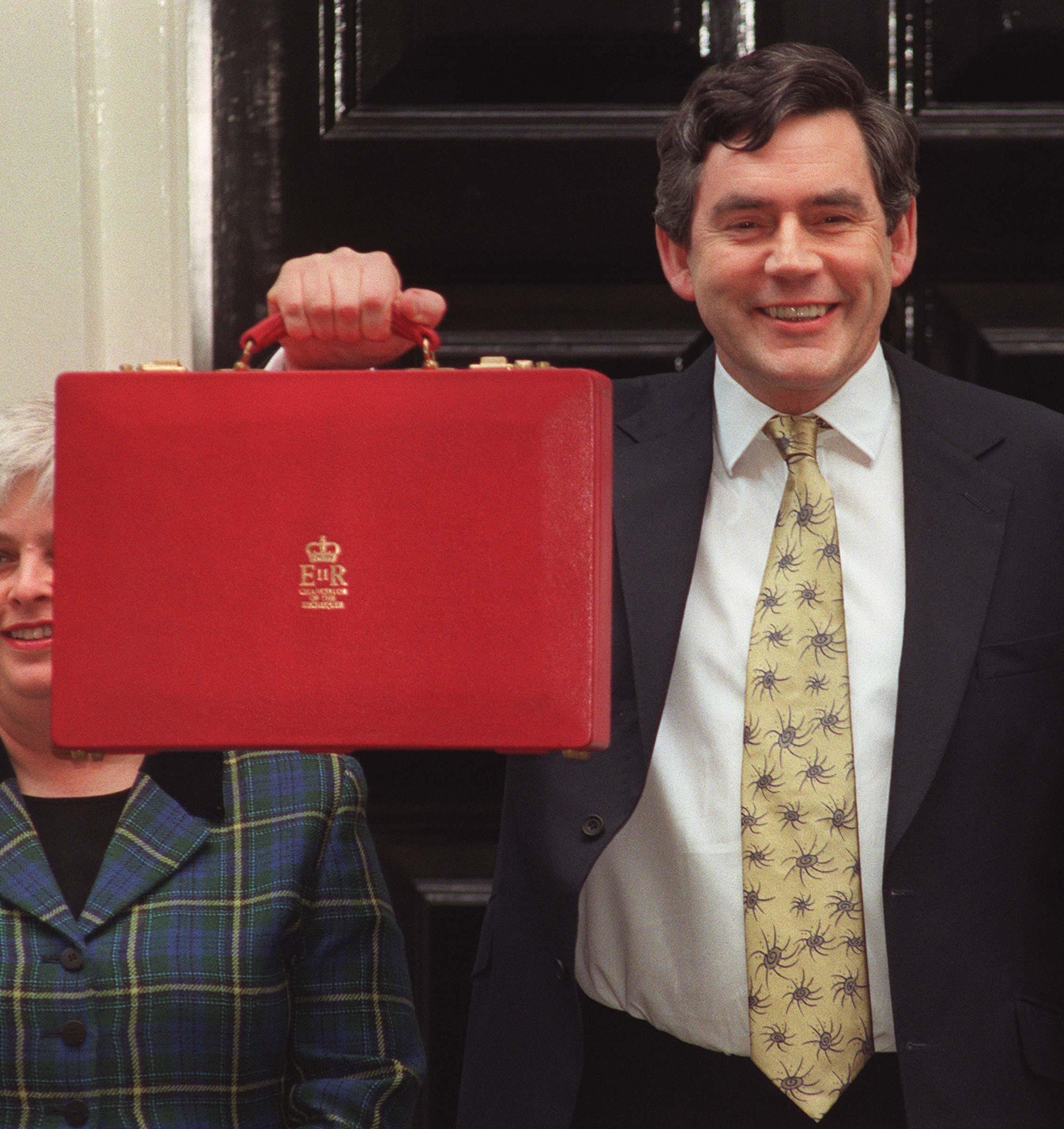 Photograph of Gordon Brown smiling and holding the red Budget briefcase outside a doorway, illustrating his role as Chancellor of the Exchequer.