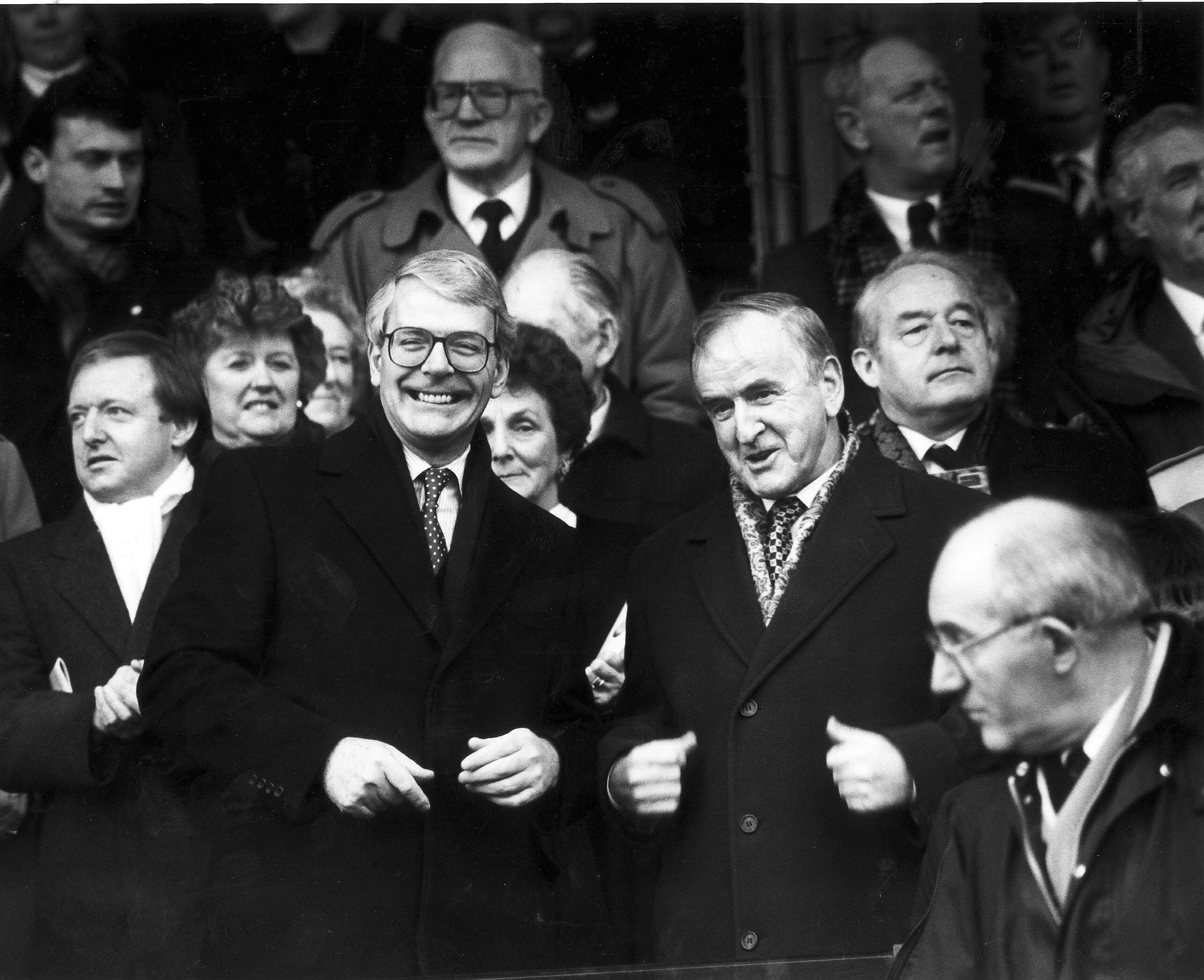 Black-and-white photograph of John Major, smiling and wearing glasses, tie and overcoat, seated among a crowded group of Conservative politicians and supporters.