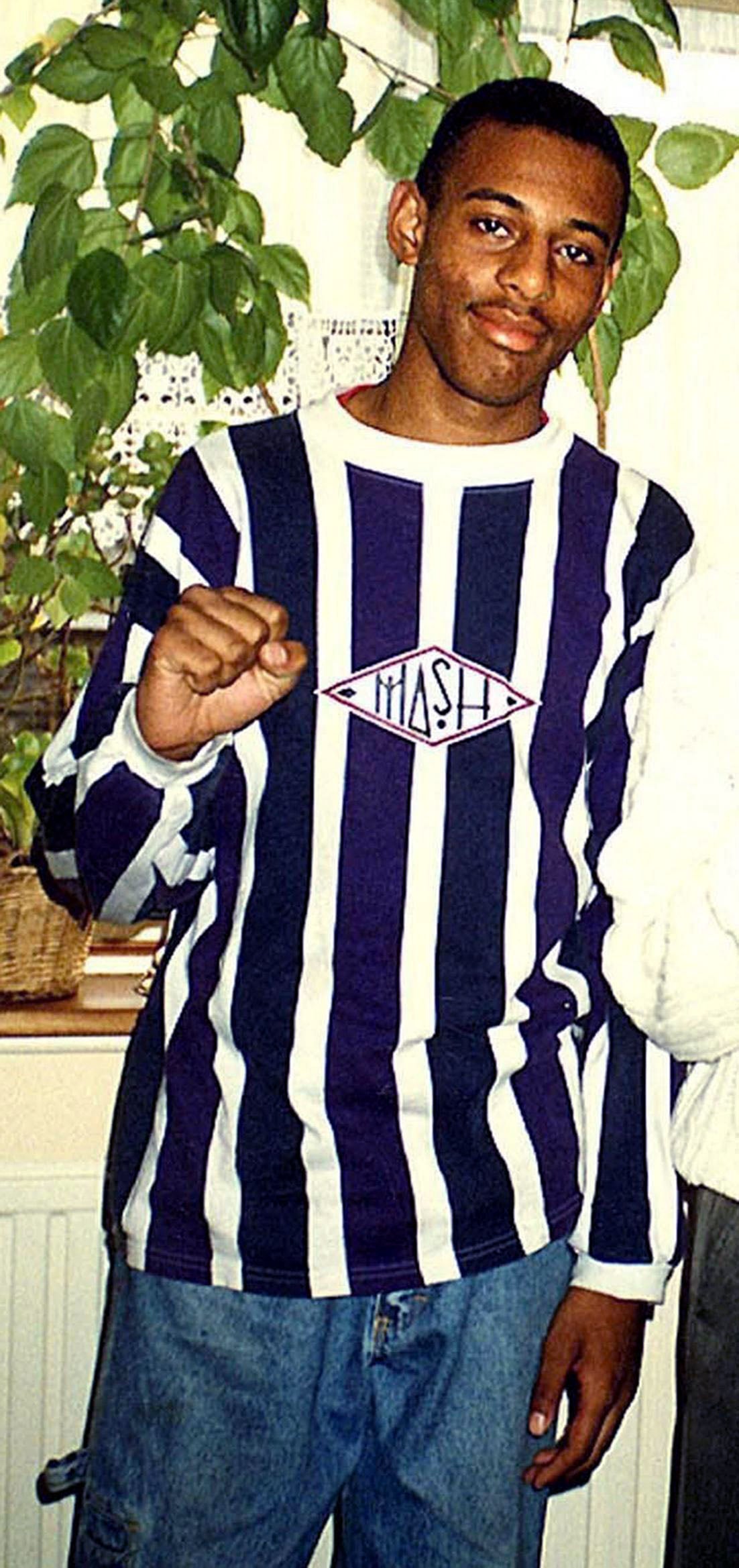 Portrait photograph of Stephen Lawrence, a young Black British man smiling at the camera in a navy-and-white striped jumper and jeans, with one hand clenched in front of him.