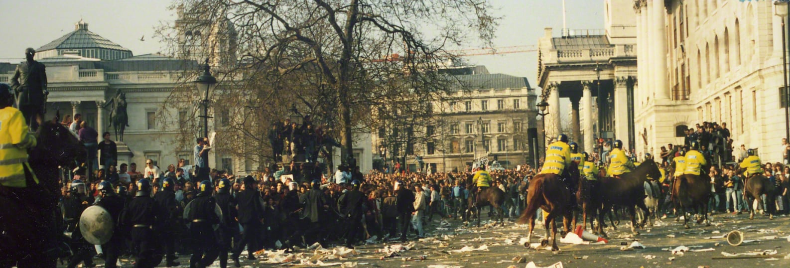 Photograph of the 31 March 1990 Trafalgar Square anti-poll-tax demonstration: riot police face a dense crowd while mounted police move through the square, with debris scattered across the street.