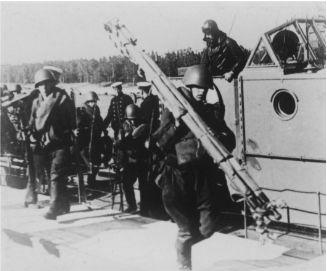 Soldiers in uniforms and helmets stand on a military boat. One soldier carries a large piece of equipment. Trees are visible in the background.