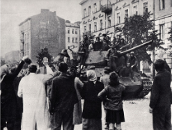 People surround a military tank on a city street, with soldiers atop it. Nearby buildings display European architecture. An air of tension is palpable.