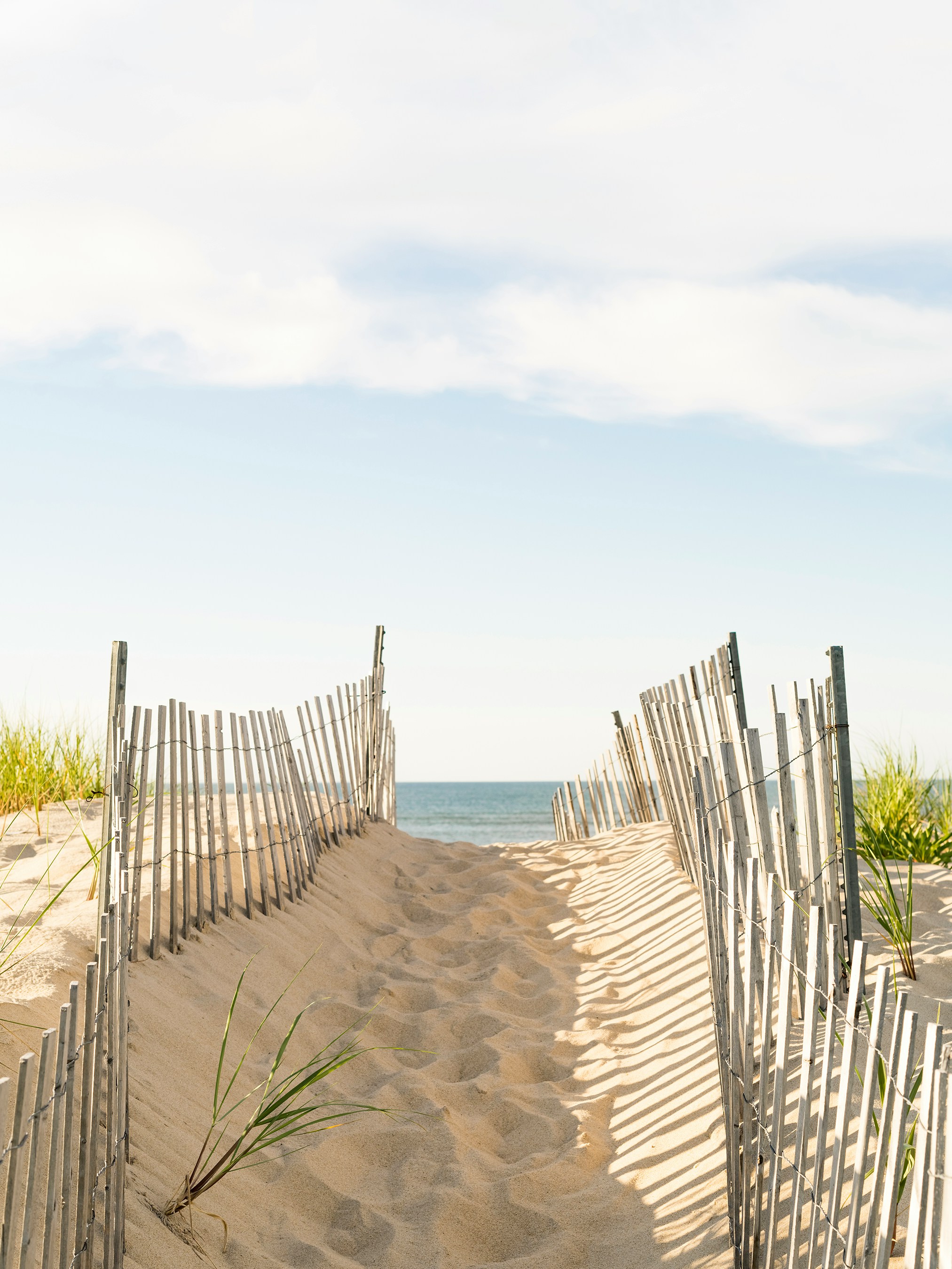 Sandy path with wooden fences leads to the sea, bordered by grass on dunes under a clear blue sky with scattered clouds.
