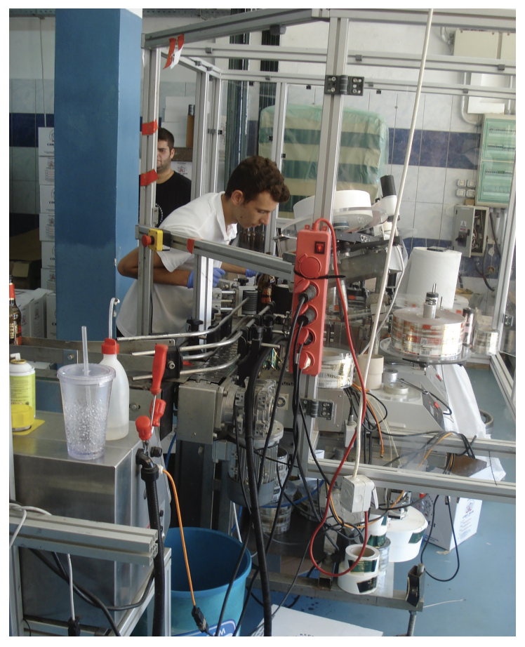 Man operating machinery with wires and bottles in an industrial setting, surrounded by tools, papers, and a blue bucket.