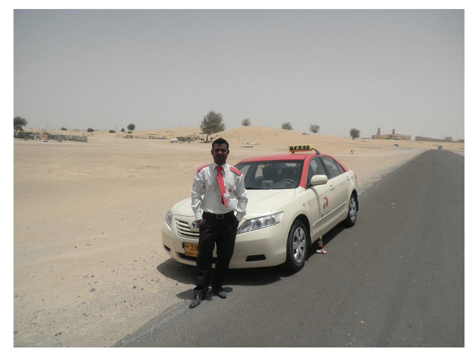 Man in white shirt and red tie stands beside a beige taxi with a red roof, parked on a desert road under a clear sky. Desert and trees in background.