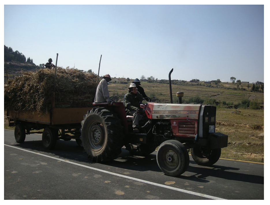 A tractor pulling a trailer filled with hay drives along a rural road, with two people seated on the tractor and fields in the background.